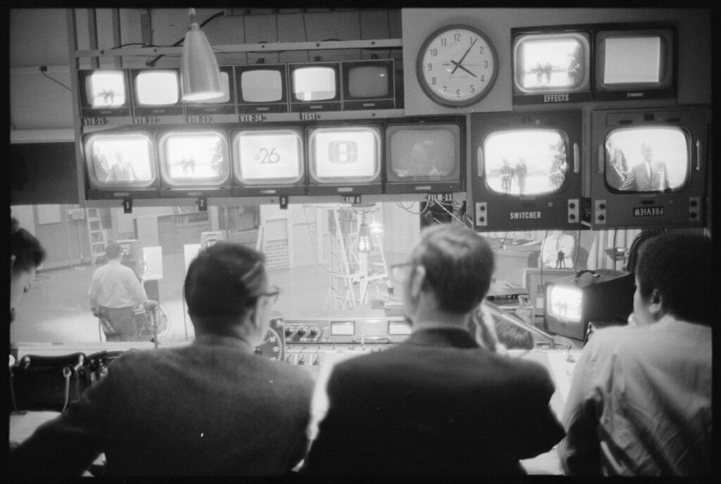 Black and white photo of people watching multiple television screens in a studio, showcasing the impact of broadcasting on viewership.