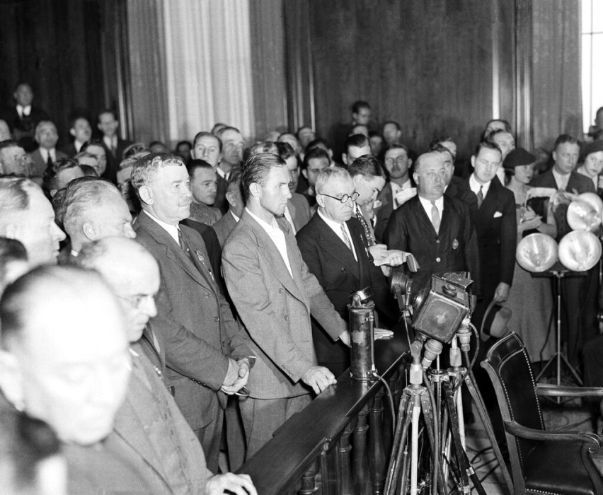 alt text: Bruno Richard Hauptmann arraignment, 1934.  Photographers with flash bulbs surround Hauptmann, highlighting the intense media presence at the time.